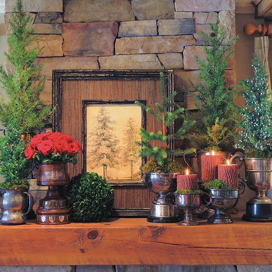 A red and green mantlescape is elevated with evergreen elements, antique trophies and a pop of bright red roses. Bark-inspired candles complete the rustic look.